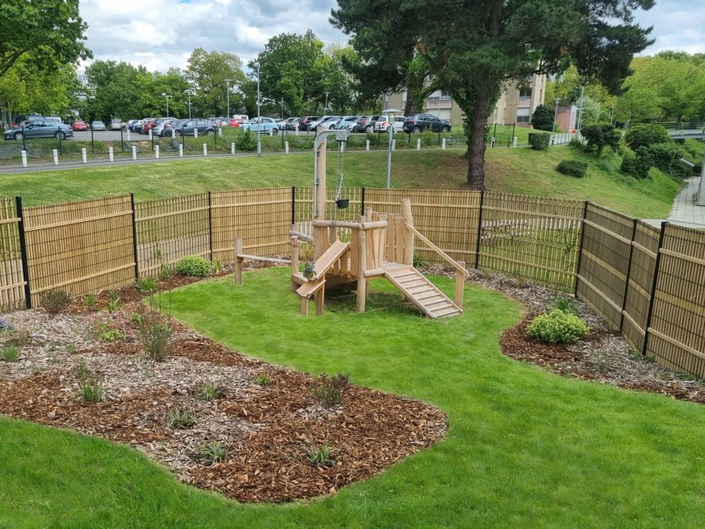 Jeu enfant en bois dans un espace enherbé et végétalisé, bordé de clôtures en panneaux rigides et occultation bois.
